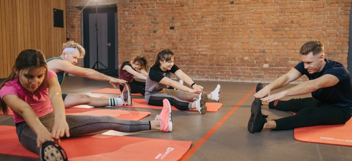 A group of diverse adults stretching on mats in a brick-walled fitness studio.